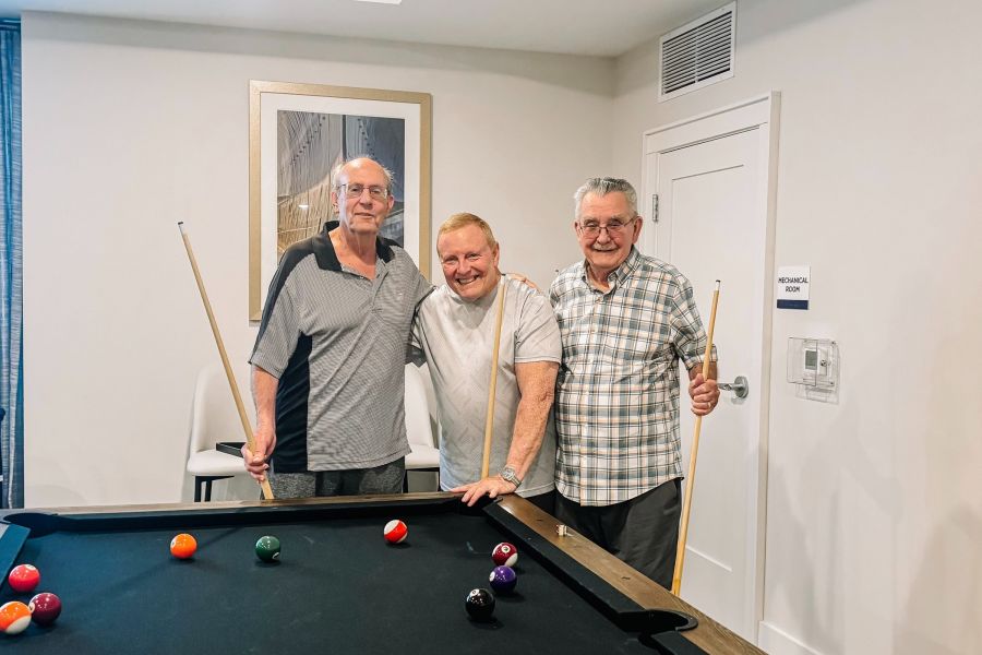 Attain at Greenbrier  apartment homes with Three older men smiling and standing by a pool table with cues, ready to play billiards in a bright room.