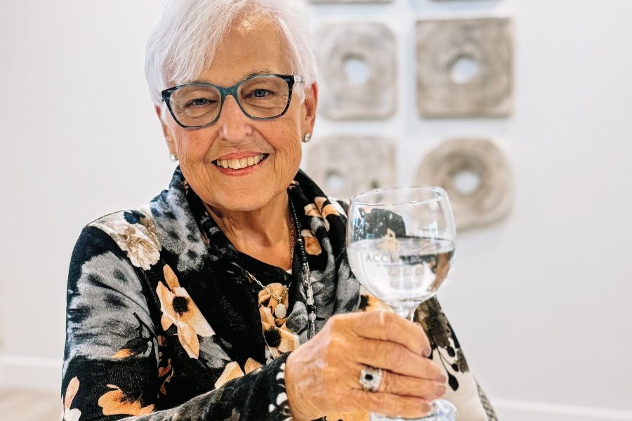 Attain at Greenbrier apartment homes with Smiling elderly woman with glasses holds up a clear glass, sitting at a table in a modern, bright room.