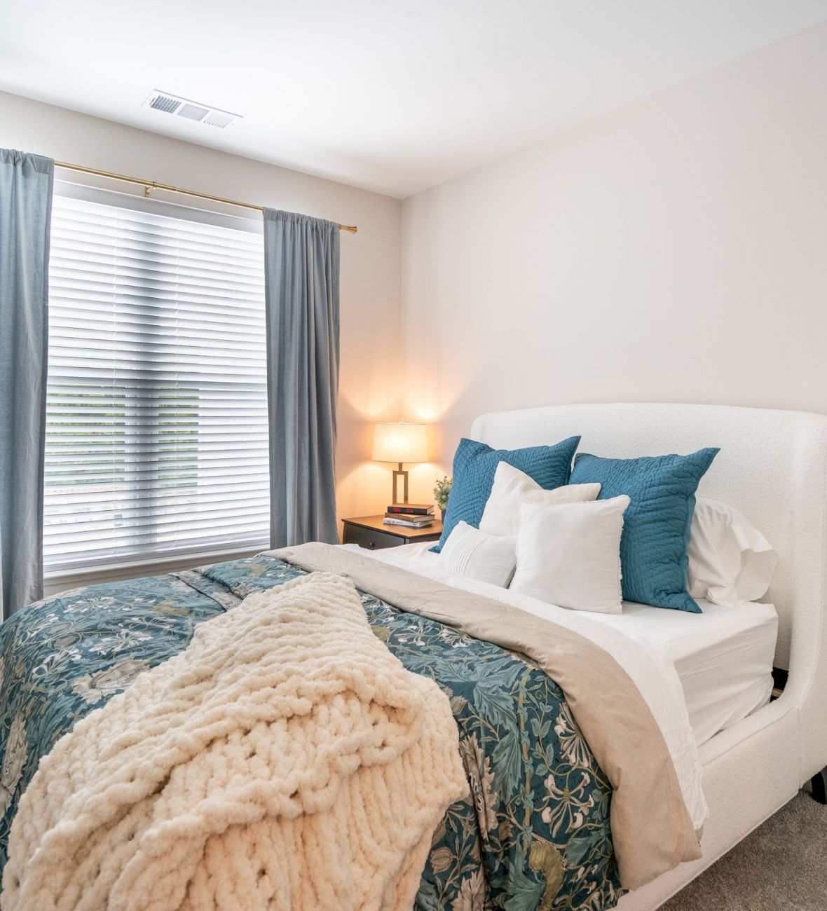 Acclaim at Greenbrier  apartment homes with Cozy bedroom with blue curtains, a bed with teal and white bedding, throw blanket, and a lit bedside lamp.
