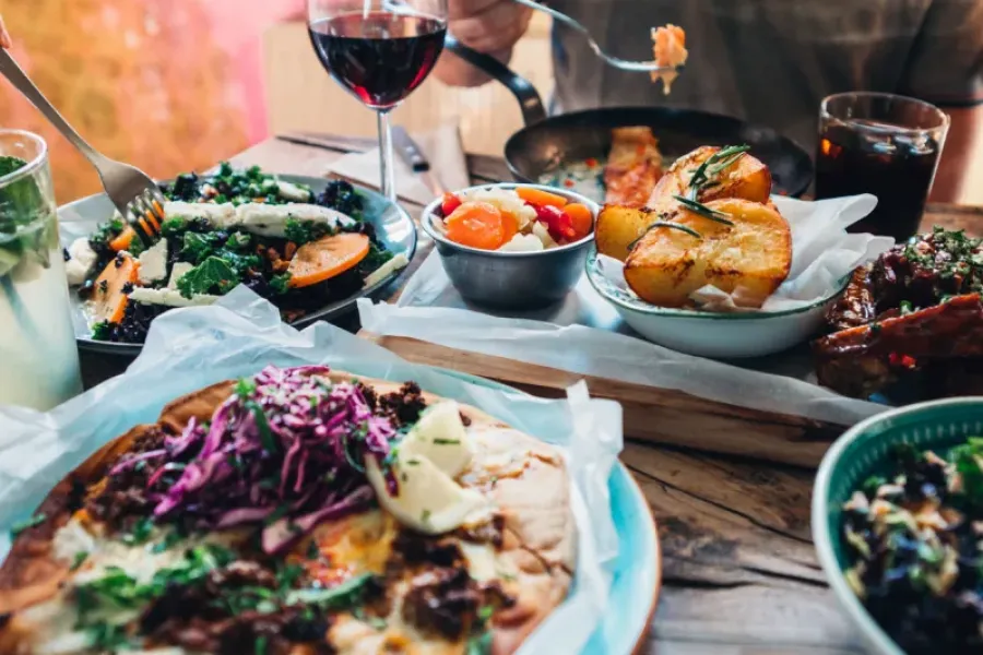 A table with pizza, salads, roasted potatoes, wine, and a person eating, all on rustic wooden table.