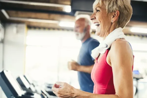 Smiling older woman jogging on a treadmill at a gym, with an older man running beside her.