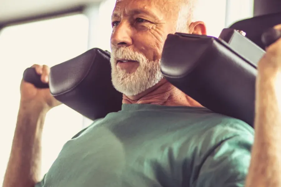 Older man with a beard using a weight machine in a gym, wearing a green shirt and smiling slightly.